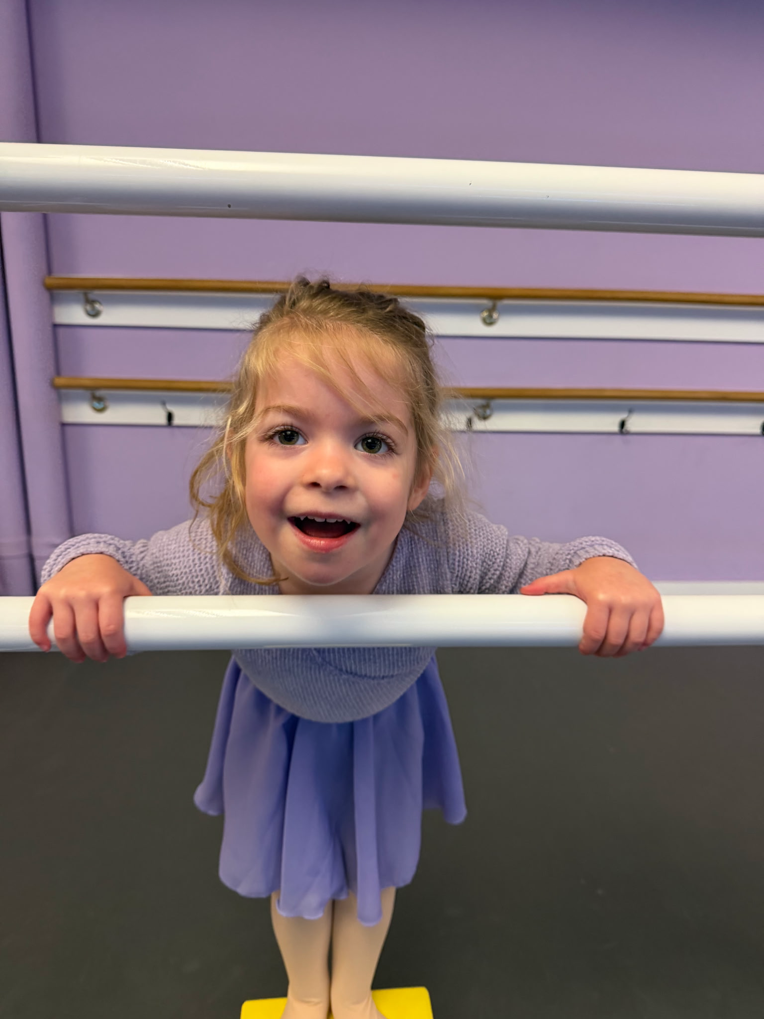A joyful preschool dancer in a lavender skirt holding the ballet barre